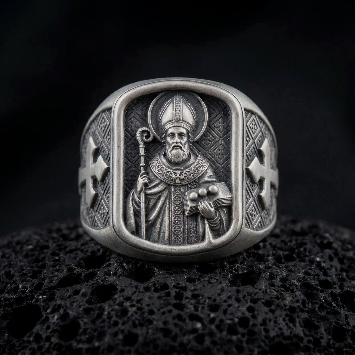 Oxidized 925 sterling silver Saint Nicholas of Myra signet ring displayed on textured black volcanic rock, featuring a bearded bishop with a mitre, crozier, book with three symbolic balls, ornate vestments, and dark oxidized silver finish.