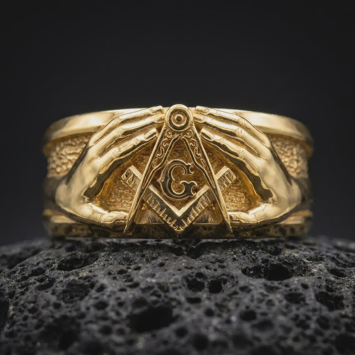 Gold plated 925 sterling silver Freemason Square and Compasses ring displayed on black volcanic rock, featuring two realistic hands surrounding the central Masonic G symbol, engraved compass arms, textured background, and warm gold plated finish.