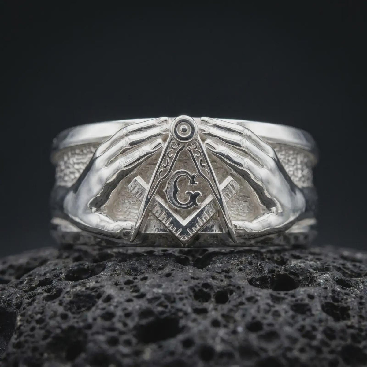 Polished 925 sterling silver Masonic ring resting on black volcanic stone, showcasing symmetrical sculpted hands surrounding a raised square and compass emblem, decorative letter G, detailed fingers, wide band, and bright polished silver finish.