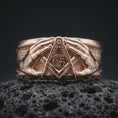 Load image into Gallery viewer, Rose gold plated 925 sterling silver Masonic hands ring resting on textured black volcanic stone, highlighting the raised Square and Compasses symbol, central letter G, symmetrical hand sculptures, decorative geometric details, and rose gold plated finish.
