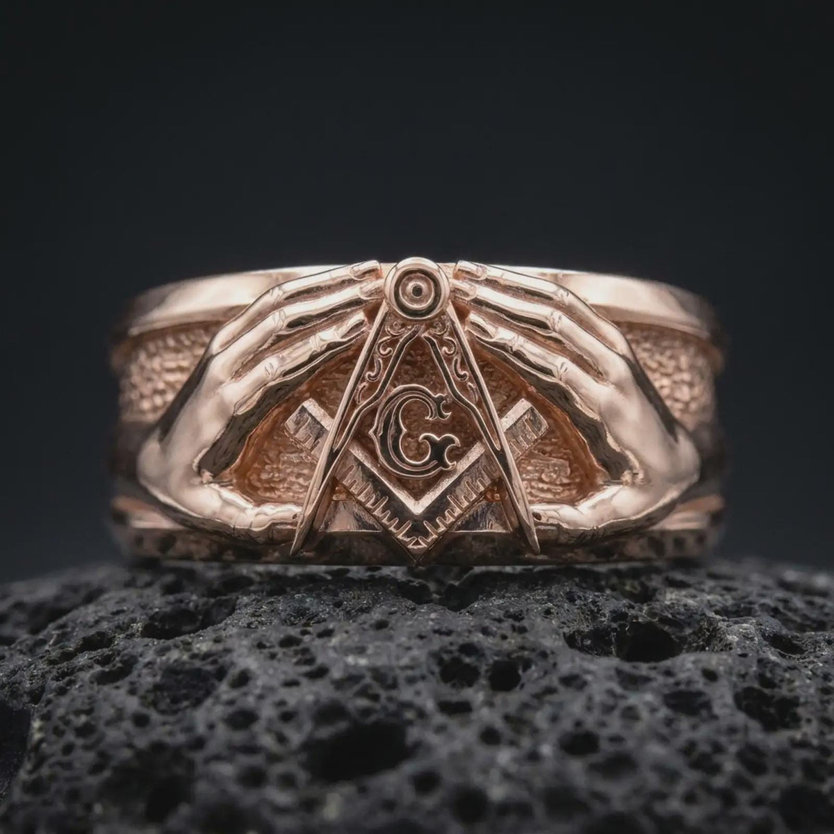 Rose gold plated 925 sterling silver Masonic hands ring resting on textured black volcanic stone, highlighting the raised Square and Compasses symbol, central letter G, symmetrical hand sculptures, decorative geometric details, and rose gold plated finish.