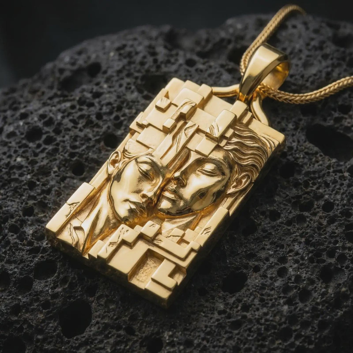 Gold plated Gemini twins pendant resting on black volcanic rock, featuring two closely positioned human faces, raised geometric patterns, and a distinctive rectangular astrology medallion.