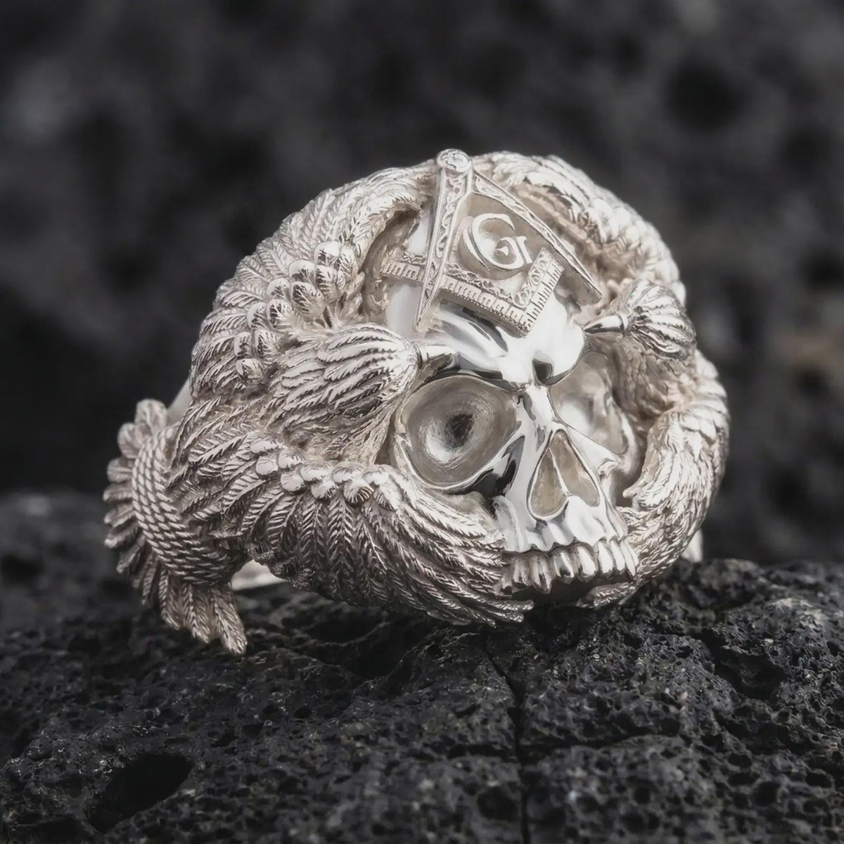 Polished sterling silver Freemason skull ring displayed on dark volcanic stone, highlighting the three-dimensional skull face, deep eye sockets, detailed teeth, Masonic forehead symbol, and layered eagle feathers.