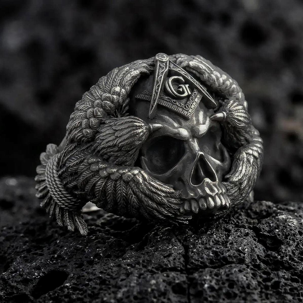 Oxidized sterling silver Masonic skull ring on black volcanic rock, featuring a detailed skull, Freemason Square and Compasses emblem with the letter G, and intricately sculpted eagle wings.