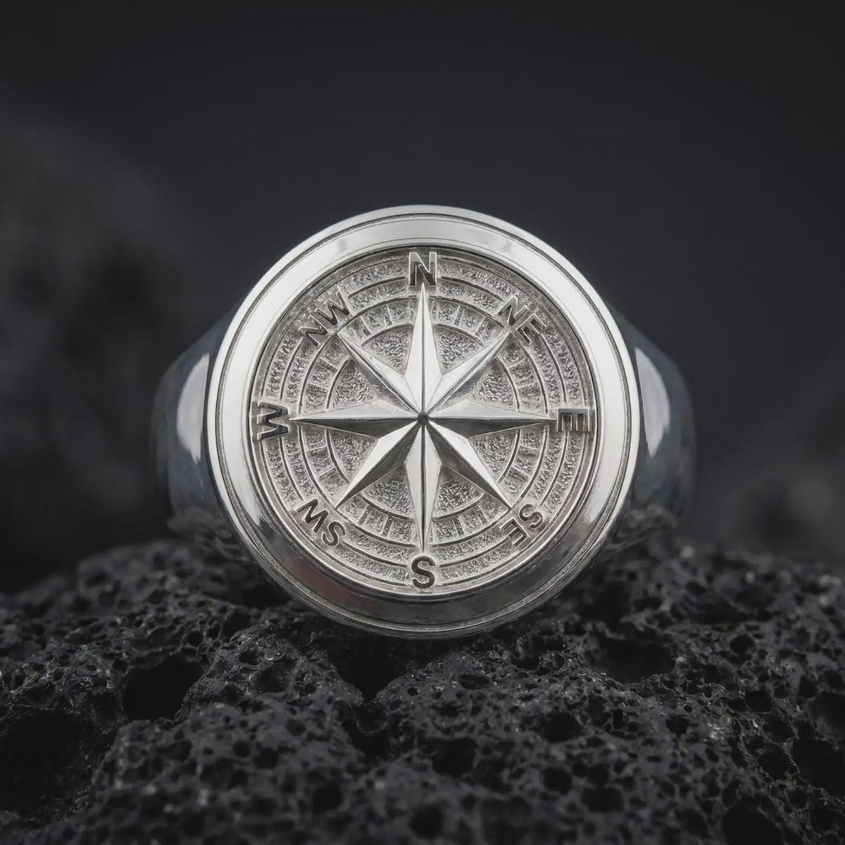 Polished sterling silver nautical compass ring displayed on dark volcanic stone, highlighting the dimensional eight-point star, cardinal direction letters, concentric compass circles, and substantial round signet face.