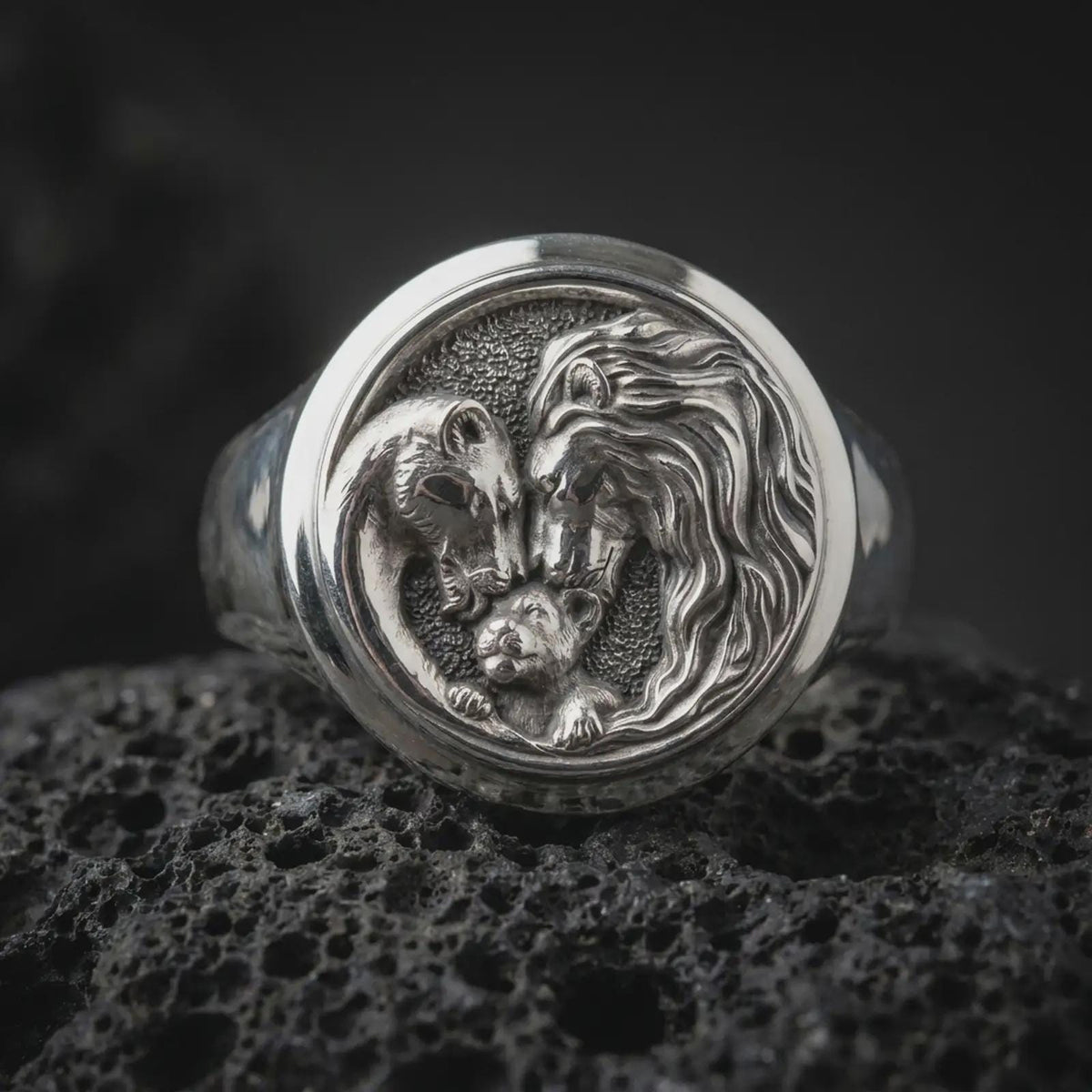 Polished sterling silver lion family ring displayed on dark volcanic stone, highlighting the detailed lion mane, gentle lioness portrait, small lion cub, and textured round signet face.