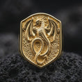 Load image into Gallery viewer, Gold plated dragon shield ring resting on black volcanic rock, featuring a mythical dragon with an elongated serpentine body, curled tail, sculpted horns, and detailed wings inside a bold heraldic shield.
