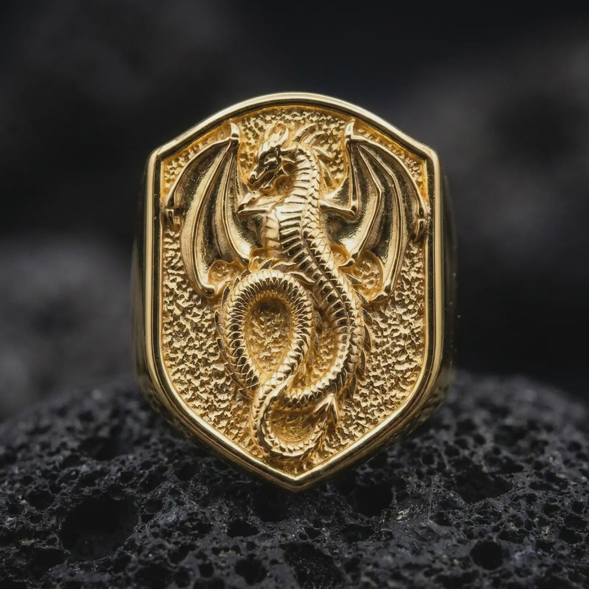 Gold plated dragon shield ring resting on black volcanic rock, featuring a mythical dragon with an elongated serpentine body, curled tail, sculpted horns, and detailed wings inside a bold heraldic shield.