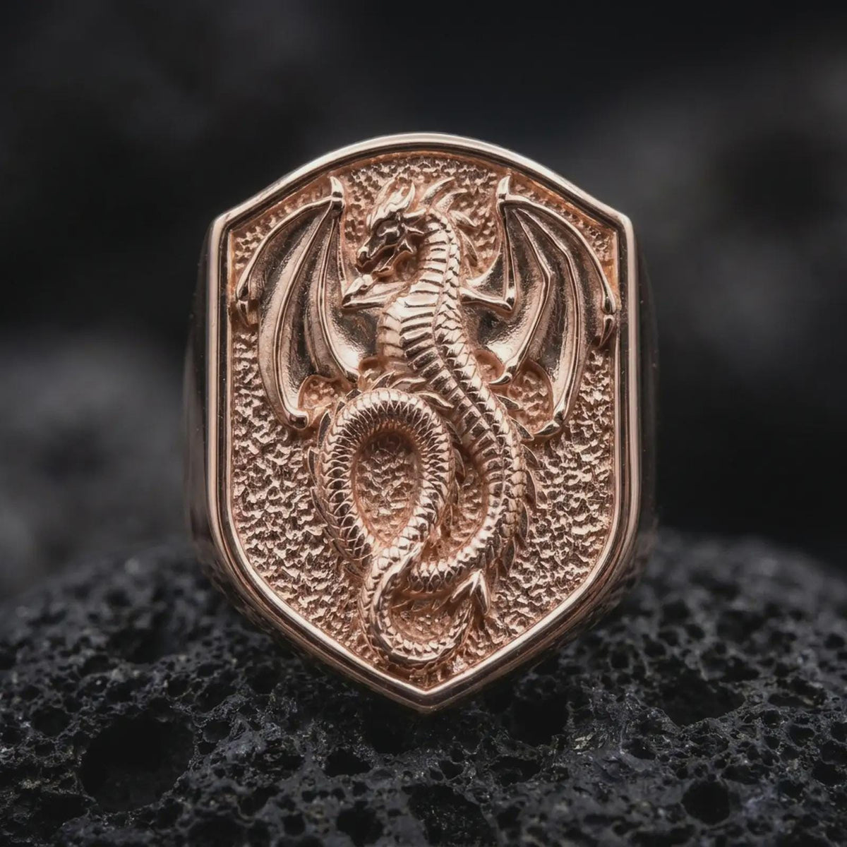 Rose gold plated medieval dragon signet ring displayed on textured volcanic stone, showcasing the raised dragon figure, detailed reptilian scales, extended wings, and distinctive shield-shaped signet face.