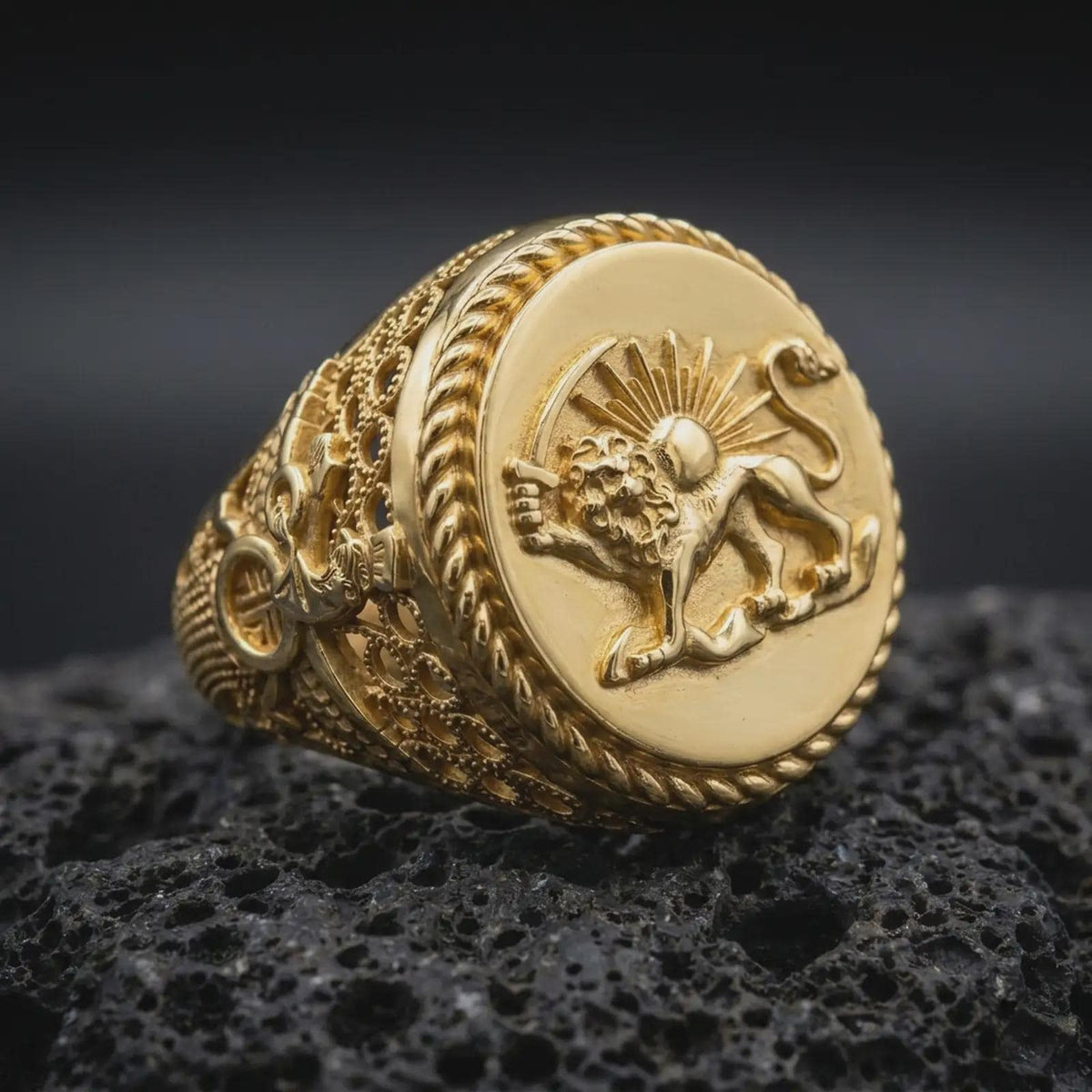 Gold plated Shir o Khorshid signet ring resting on black volcanic rock, featuring the historic Iranian lion and sun symbol, raised sword-bearing lion, radiant sun, and detailed circular rope-edge frame.