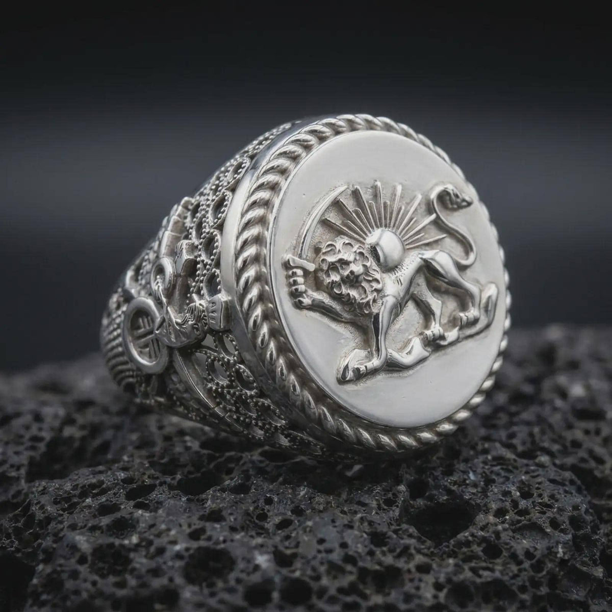 Polished sterling silver Persian Lion and Sun ring displayed on dark volcanic stone, highlighting the sculpted lion relief, sunburst rays, antique coin-style face, and intricate decorative band details.