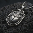 Load image into Gallery viewer, Oxidized sterling silver stag beetle shield pendant on black volcanic rock, featuring a raised beetle with antler-like mandibles, detailed wing covers, sculpted legs, and a textured shield background.
