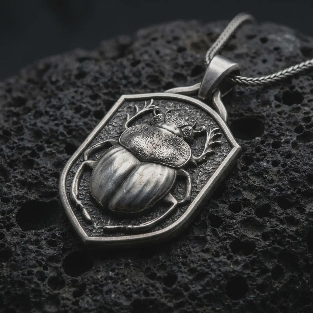 Oxidized sterling silver stag beetle shield pendant on black volcanic rock, featuring a raised beetle with antler-like mandibles, detailed wing covers, sculpted legs, and a textured shield background.