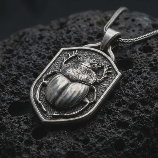 Oxidized sterling silver stag beetle shield pendant on black volcanic rock, featuring a raised beetle with antler-like mandibles, detailed wing covers, sculpted legs, and a textured shield background.