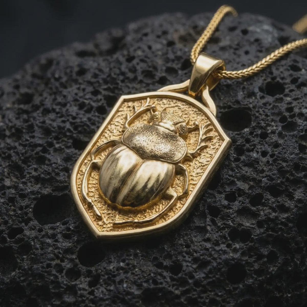 Gold plated stag beetle pendant resting on black volcanic rock, featuring a sculpted insect with prominent antler-like jaws, rounded armored wing covers, and detailed legs against a medieval-style shield.