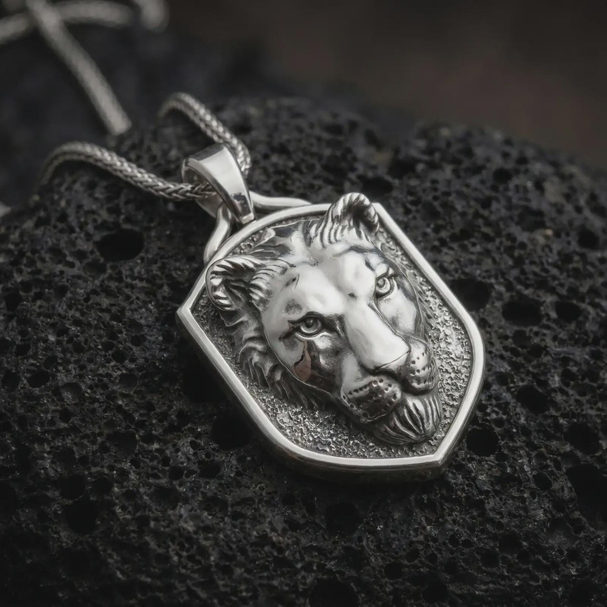 Polished sterling silver lioness pendant displayed on dark volcanic rock, highlighting the sculpted wild cat face, focused eyes, defined ears, smooth muzzle, and bold shield-shaped silver border.