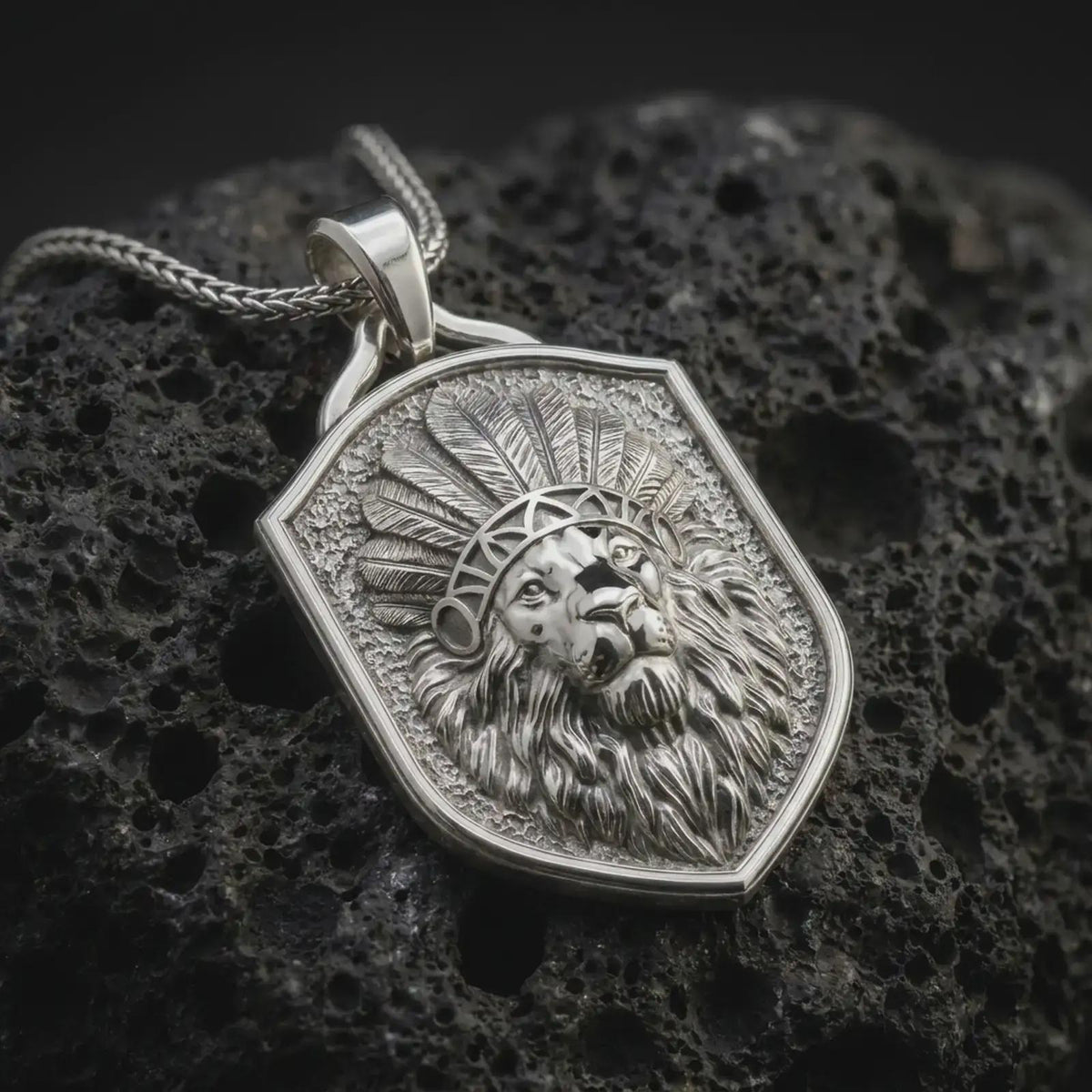Polished sterling silver lion headdress pendant displayed on dark volcanic rock, highlighting the three-dimensional lion portrait, layered ceremonial feathers, expressive eyes, detailed muzzle, and sculpted mane.