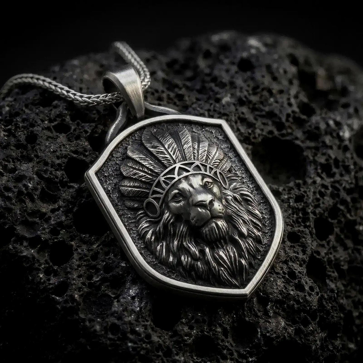 Oxidized sterling silver lion chief pendant necklace on black volcanic stone, featuring a majestic lion head wearing a detailed feather headdress, flowing mane, textured shield-shaped background, and braided silver chain.