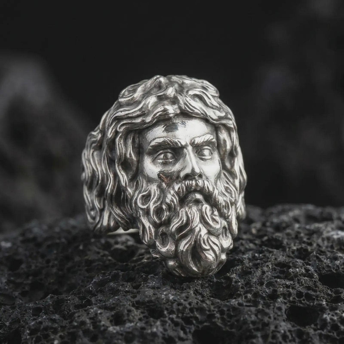 Polished 925 sterling silver Poseidon ring resting on black volcanic stone, highlighting the detailed Greek god portrait, expressive facial features, curled mustache, flowing sculptural hair, and bright polished silver finish.