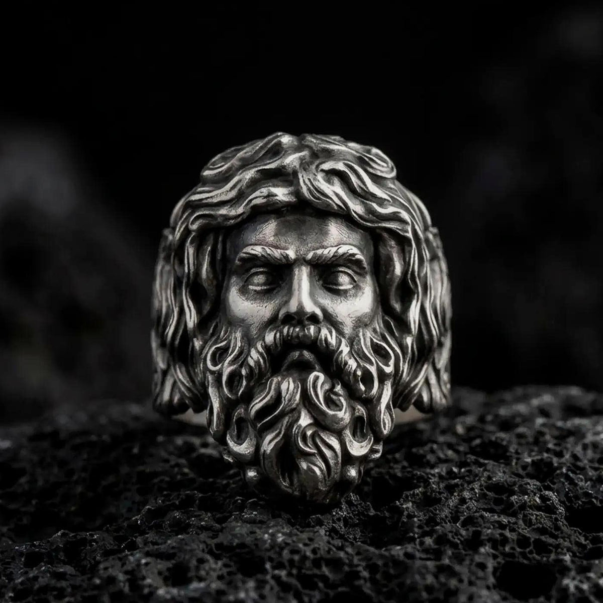 Oxidized 925 sterling silver Poseidon ring displayed on black volcanic rock, featuring a three-dimensional Greek sea god face with flowing wavy hair, deeply sculpted beard, prominent eyebrows, and dark recessed details.