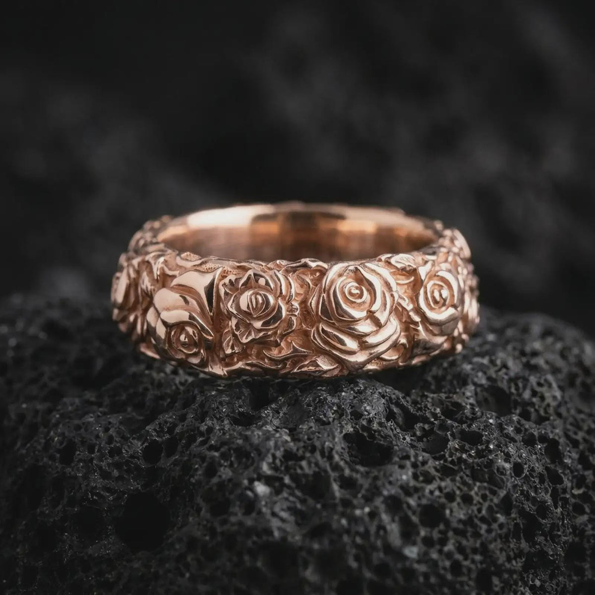 Rose gold plated sterling silver floral band ring resting on black volcanic stone, highlighting continuous sculpted rose blossoms, delicate leaves, dimensional flower petals, and a romantic rose gold finish.