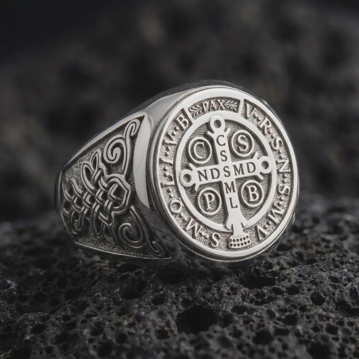 White gold Saint Benedict signet ring resting on black volcanic stone, showcasing the engraved Catholic cross medal, traditional Latin letters, PAX symbol, intricate Celtic-inspired shoulder patterns, and bright polished silver finish.