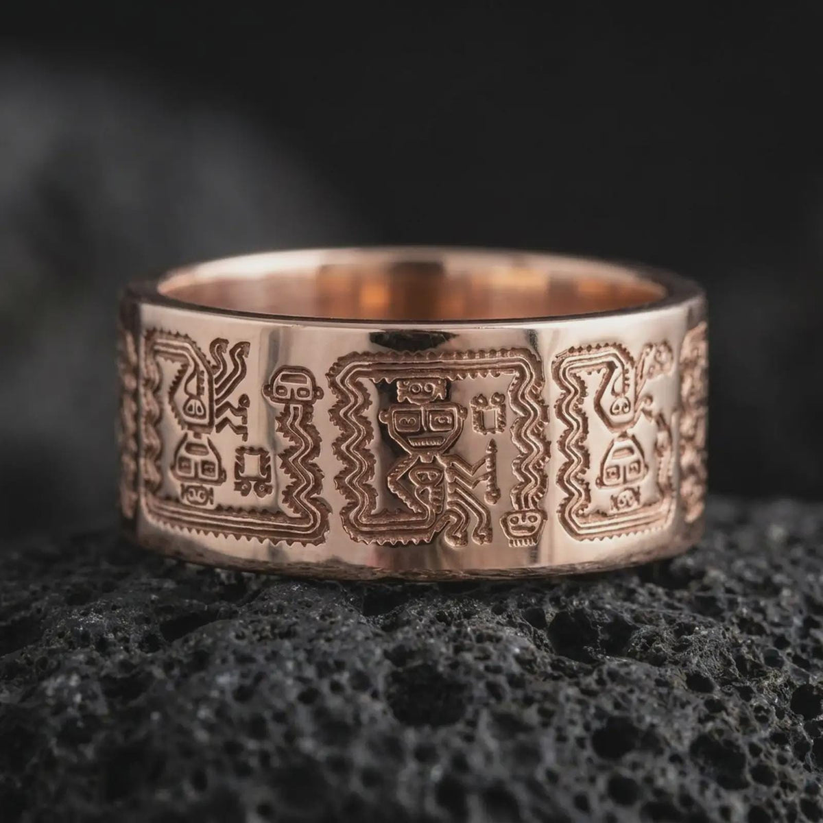 Rose gold plated 925 sterling silver Aztec Mayan band resting on black volcanic stone, highlighting intricate symbolic figures, angular geometric patterns, repeating ancient-inspired engravings, and a warm rose gold finish.
