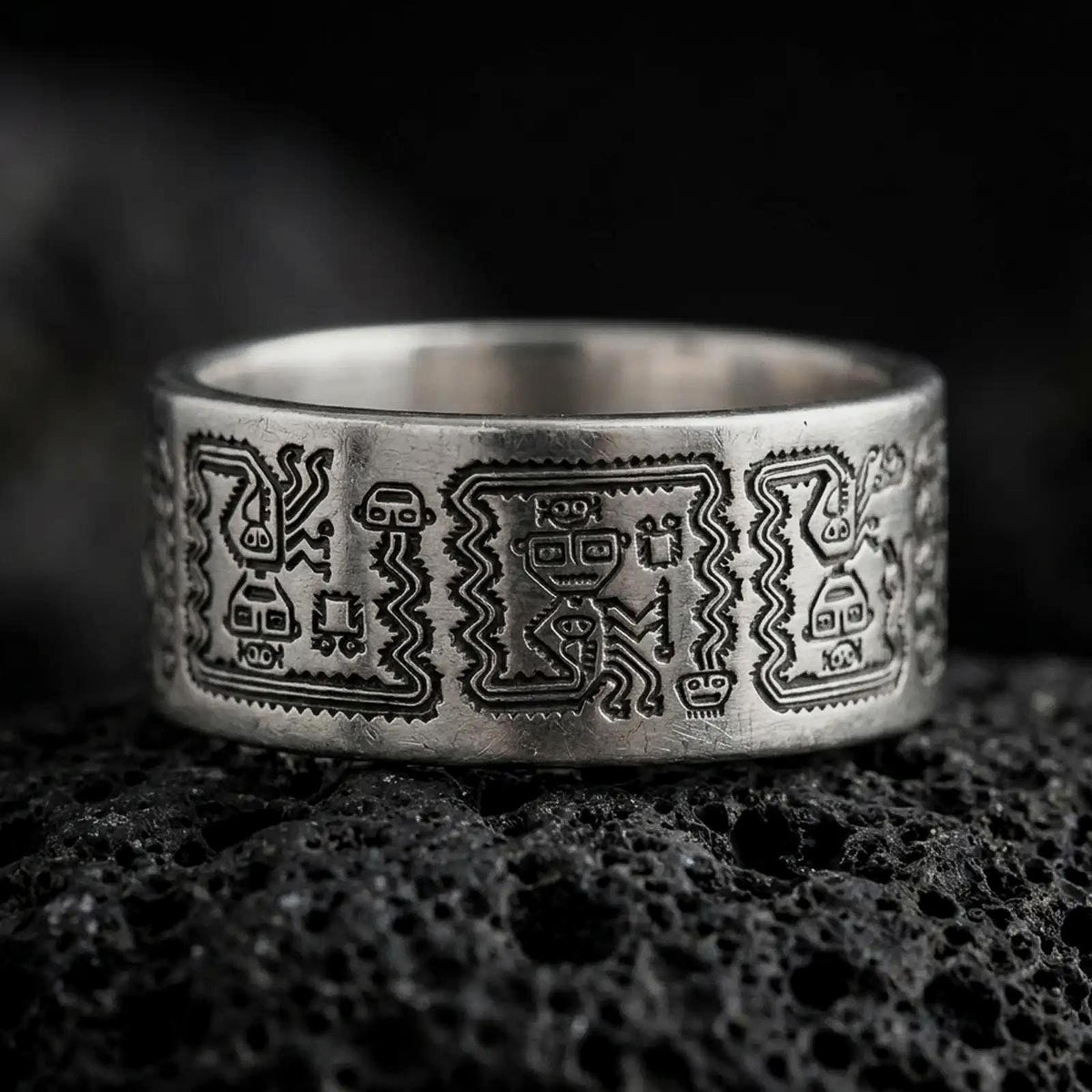 Oxidized 925 sterling silver Aztec Mayan ring displayed on black volcanic rock, featuring intricate Mesoamerican-inspired figures, stepped geometric patterns, dark recessed engravings, and a wide silver wedding band.
