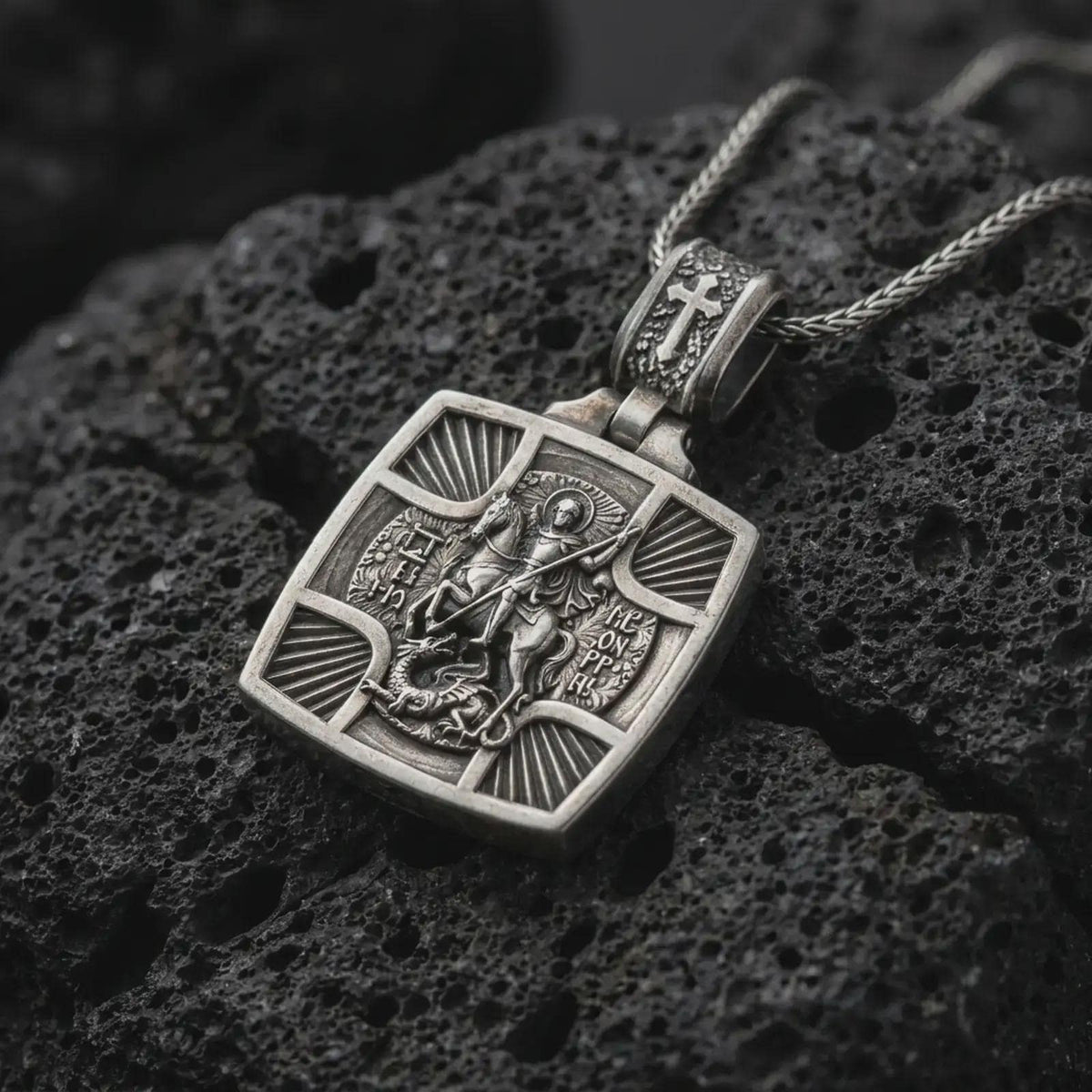 Oxidized Silver Saint George Slaying Dragon pendant on black volcanic rock, featuring the warrior saint on horseback with a spear.