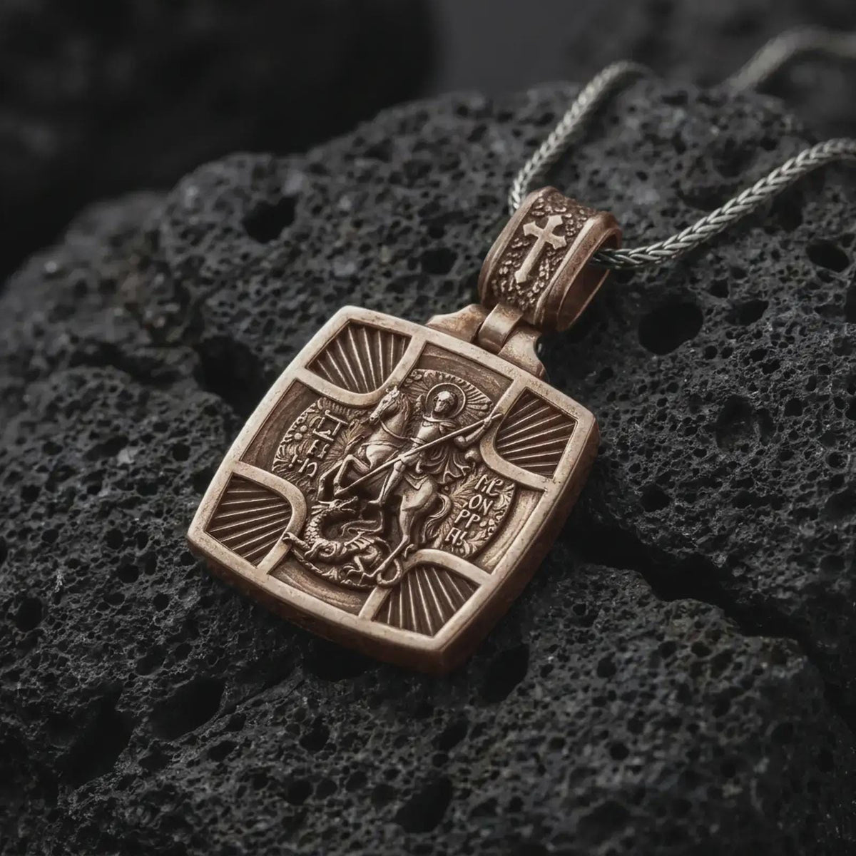 Bronze Saint George Slaying Dragon pendant on black volcanic rock, featuring the warrior saint on horseback with a spear.