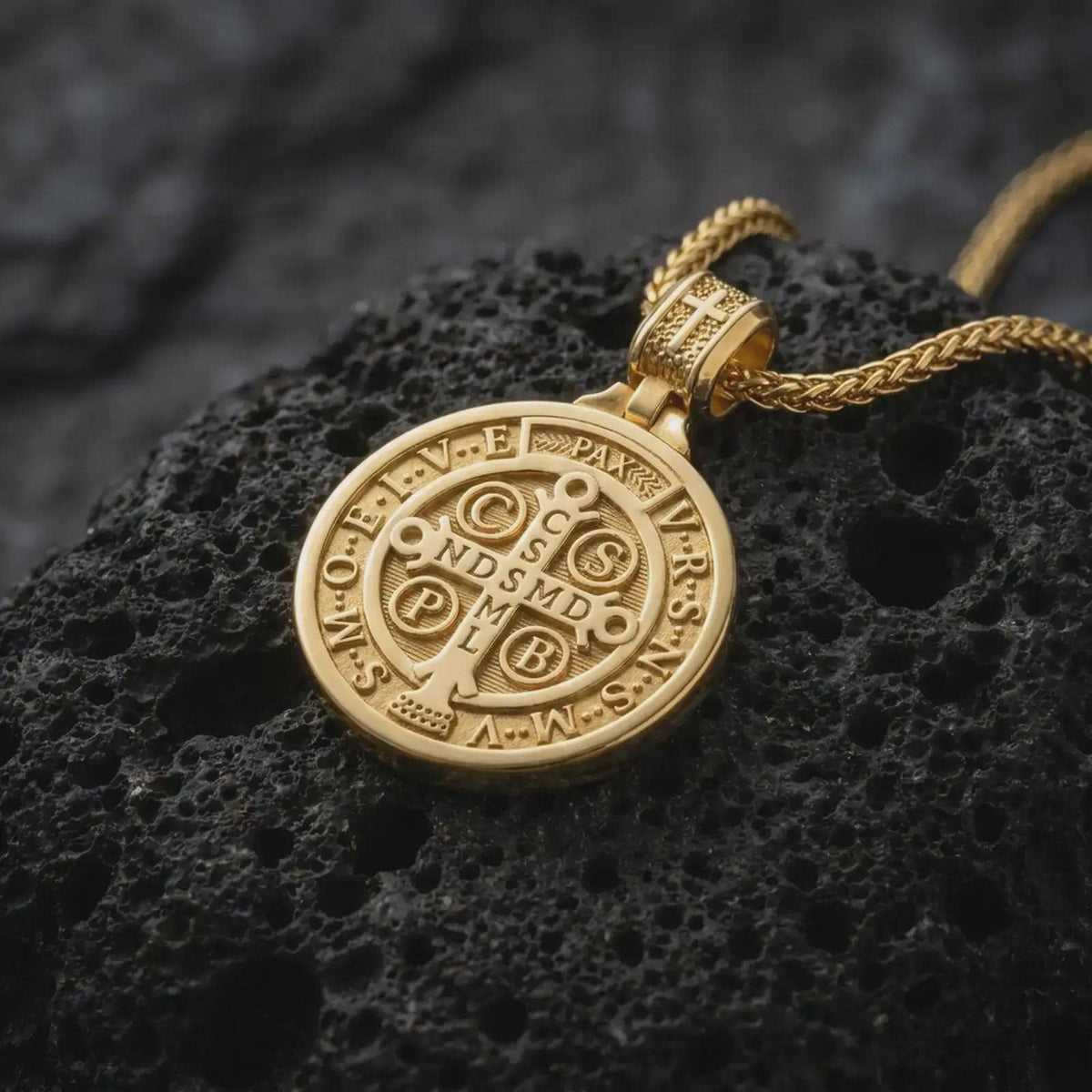 Gold Plated double sided Saint Benedict pendant displayed on black volcanic rock, highlighting the Saint Benedict medal side and cross-decorated bail.