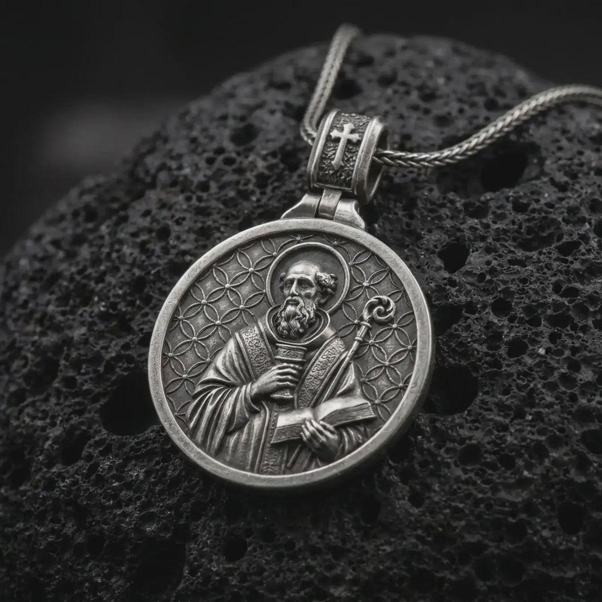 Oxidized Silver double sided Saint Benedict pendant on black volcanic rock, showing the Benedict medal cross with Latin initials and PAX inscription.