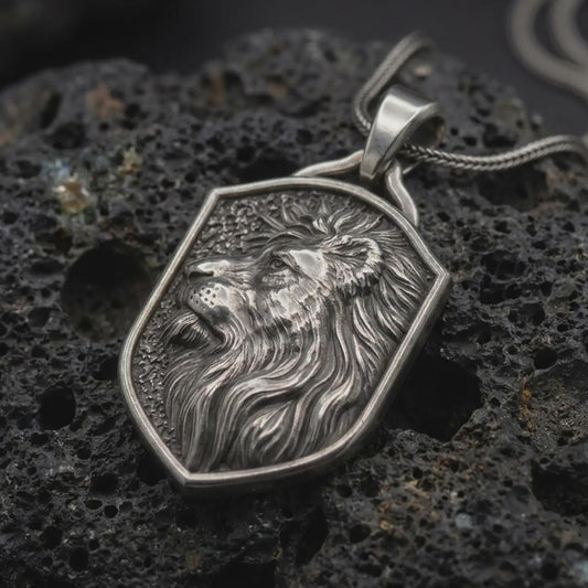 Oxidized 925 sterling silver lion head shield pendant displayed on textured black volcanic rock, featuring a majestic side-profile lion with an open mouth, deeply sculpted flowing mane, angular shield frame, dark recessed background, and oxidized silver finish.