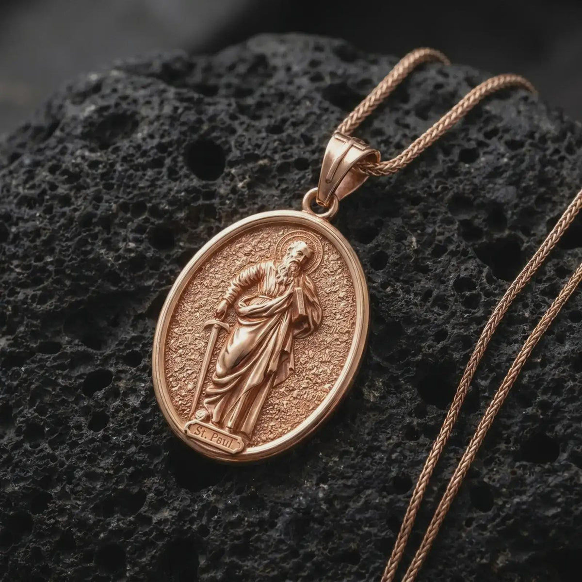 Rose Gold Plated Saint Paul Catholic medal resting on black volcanic rock, featuring the bearded apostle with his traditional book and sword.