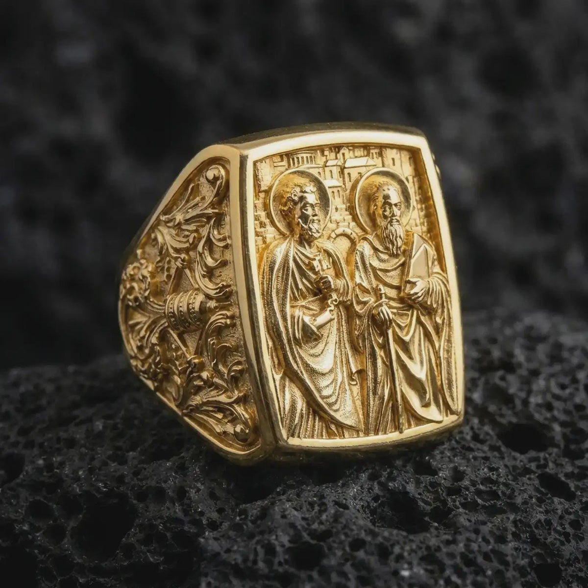 Gold Plated Saint Peter and Paul signet ring displayed on black volcanic rock, showing the sculpted apostle figures and decorative floral patterns.