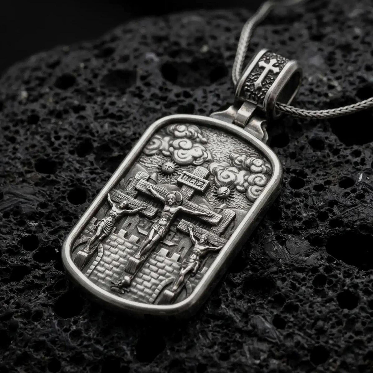 Oxidized Silver Three Crosses Crucifixion pendant on black volcanic rock, featuring Jesus Christ between two thieves at Calvary.