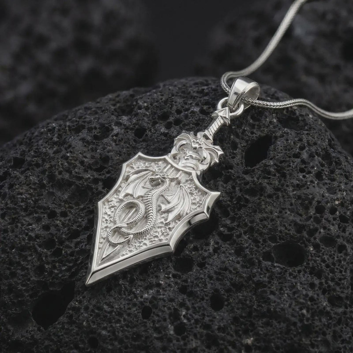 Polished 925 sterling silver dragon sword necklace resting on black volcanic stone, showcasing a detailed winged dragon with a curled tail, spread wings, sculpted scales, ornate sword crossguard, shield-shaped blade, and bright polished silver finish.