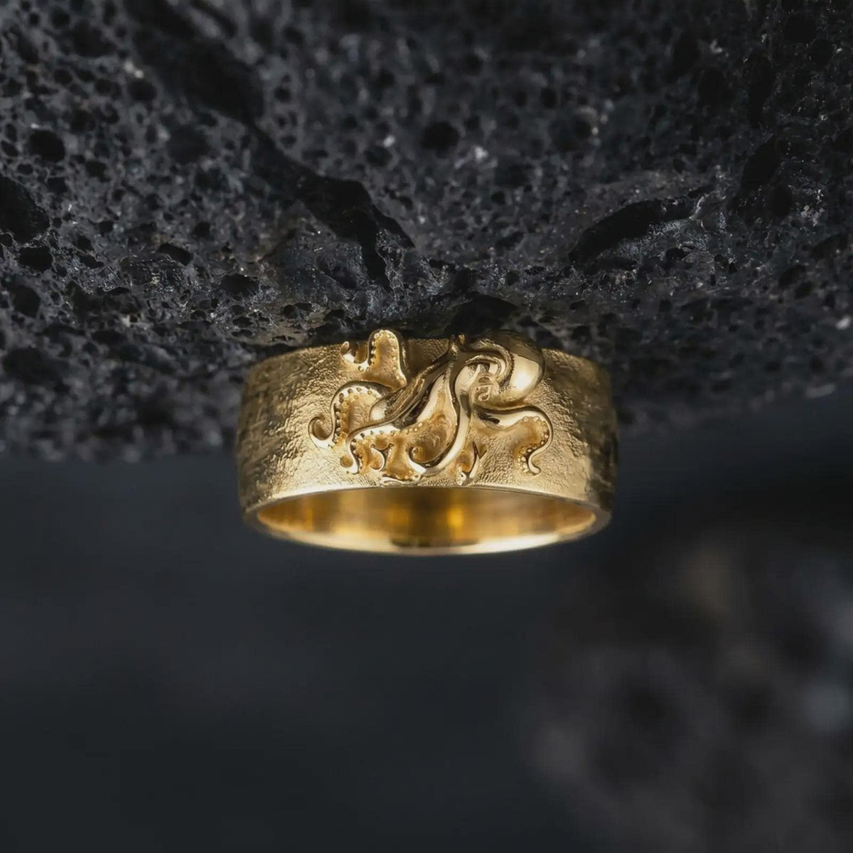 Gold plated 925 sterling silver octopus tentacle band displayed against textured black volcanic rock, featuring a raised sea creature sculpture, curling tentacles, detailed suction cups, flowing organic patterns, and warm gold plated finish.
