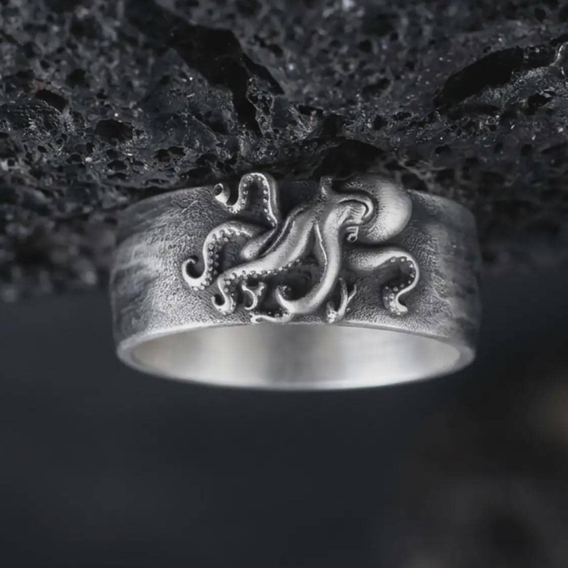 Oxidized 925 sterling silver octopus tentacle ring displayed against textured black volcanic rock, featuring a sculpted octopus with curling arms, raised suction cups, flowing tentacles, wide band design, and dark oxidized silver finish.