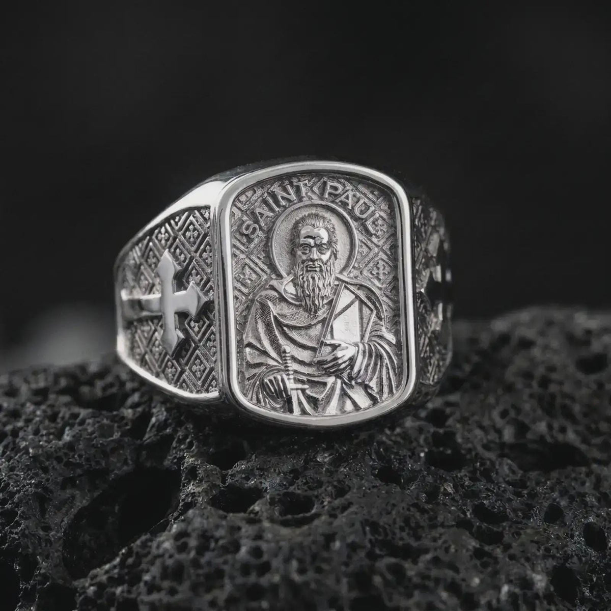 Polished Silver Saint Paul ring on black volcanic rock, highlighting the rectangular saint portrait, engraved inscription, and geometric background.