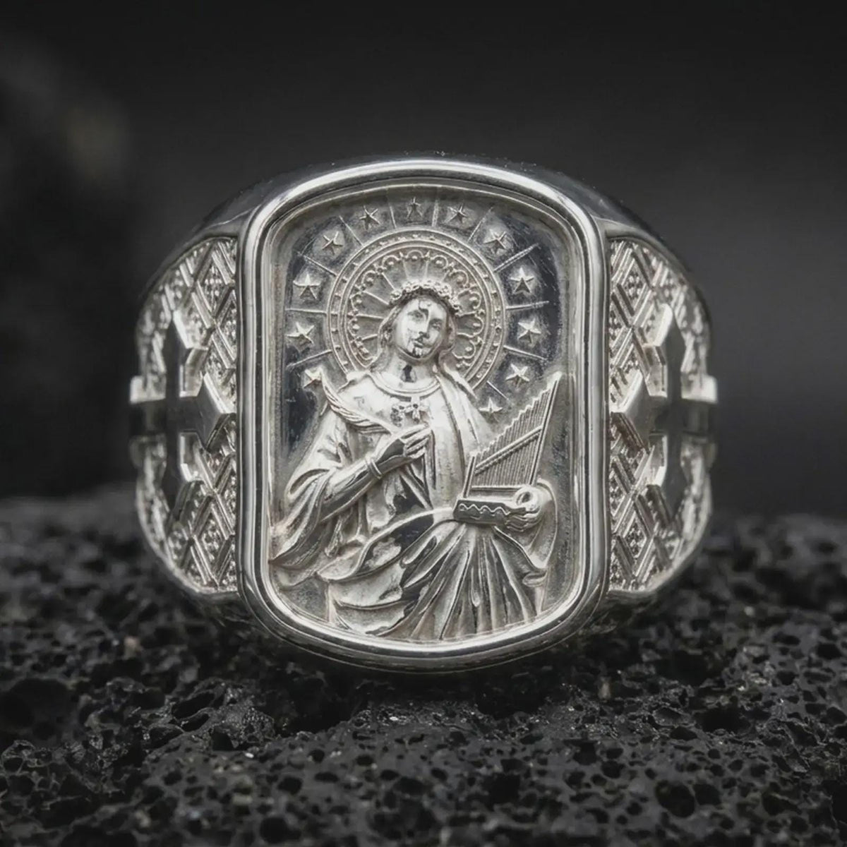 Polished Silver Saint Cecilia ring on black volcanic rock, highlighting the star-adorned halo, floral crown, and detailed sculptural relief.