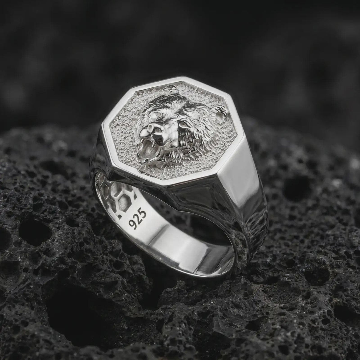 Polished 925 sterling silver bear head ring resting on black volcanic stone, showcasing a fierce roaring bear with sculpted facial fur, exposed canine teeth, textured hexagonal signet face, and bright polished silver finish.