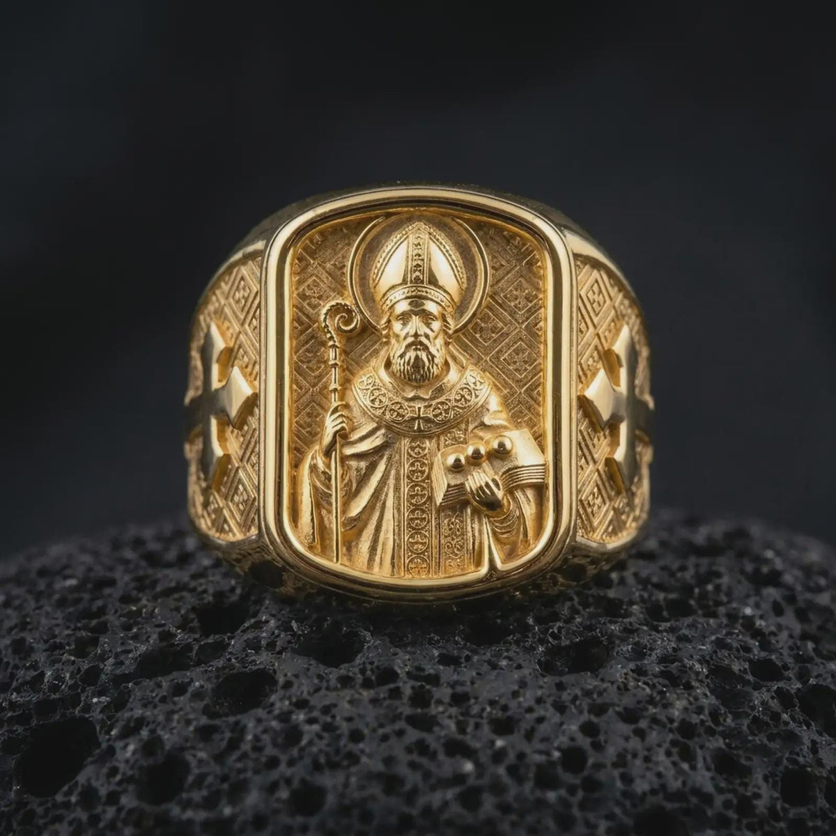 Gold plated 925 sterling silver Saint Nicholas signet ring displayed on textured black volcanic rock, featuring the Catholic bishop with detailed religious robes, bishop's crozier, book with three symbolic balls, engraved cross shoulders, and warm gold plated finish.