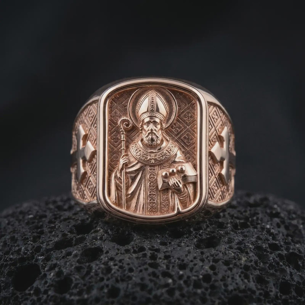 Rose gold plated 925 sterling silver Saint Nicholas of Myra ring resting on black volcanic stone, highlighting the raised bishop portrait, ornate mitre, halo, pastoral staff, symbolic three-ball book, geometric background, and warm rose gold plated finish.