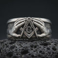 Load image into Gallery viewer, Oxidized 925 sterling silver Freemason ring displayed on textured black volcanic rock, featuring two sculpted hands framing the Masonic Square and Compasses, central letter G, dark recessed background, and oxidized silver finish.
