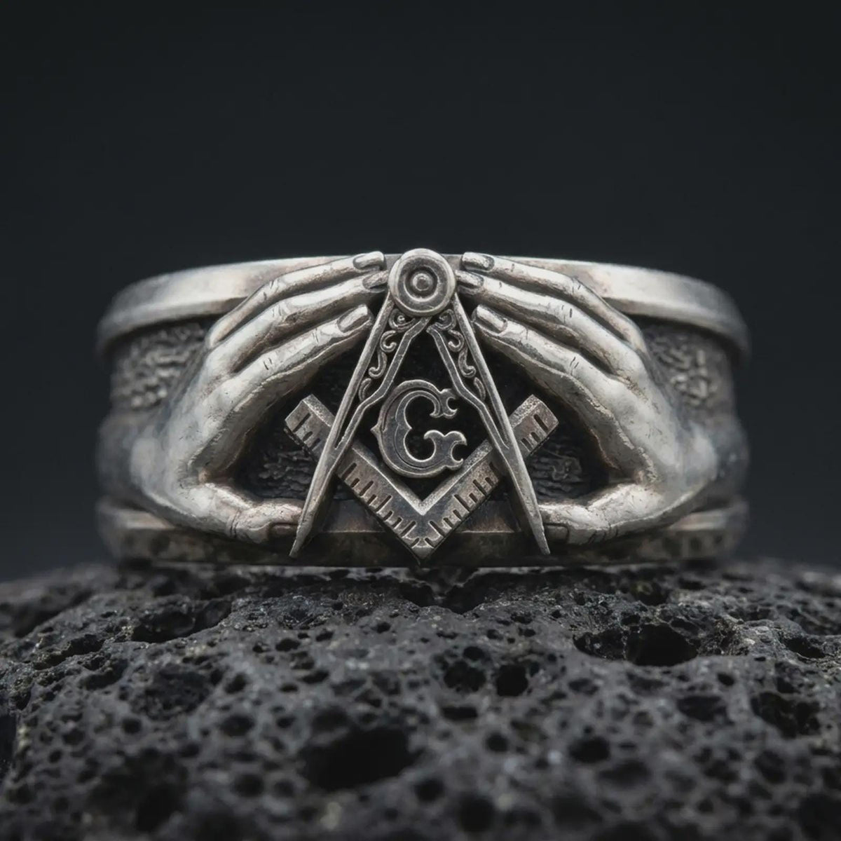 Oxidized 925 sterling silver Freemason ring displayed on textured black volcanic rock, featuring two sculpted hands framing the Masonic Square and Compasses, central letter G, dark recessed background, and oxidized silver finish.