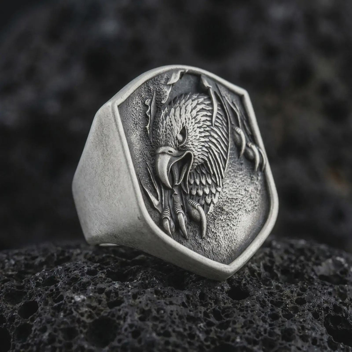 Oxidized 925 sterling silver eagle head signet ring displayed on textured black volcanic rock, featuring a screaming eagle with an open hooked beak, sharp talons, detailed feathers, shield-shaped face, and dark oxidized silver finish.