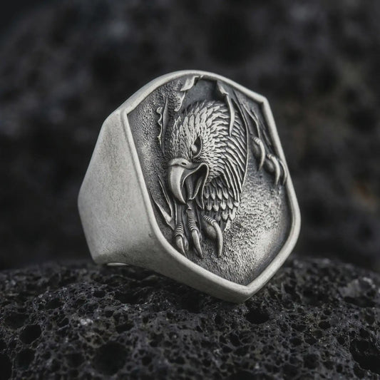 Oxidized 925 sterling silver eagle head signet ring displayed on textured black volcanic rock, featuring a screaming eagle with an open hooked beak, sharp talons, detailed feathers, shield-shaped face, and dark oxidized silver finish.