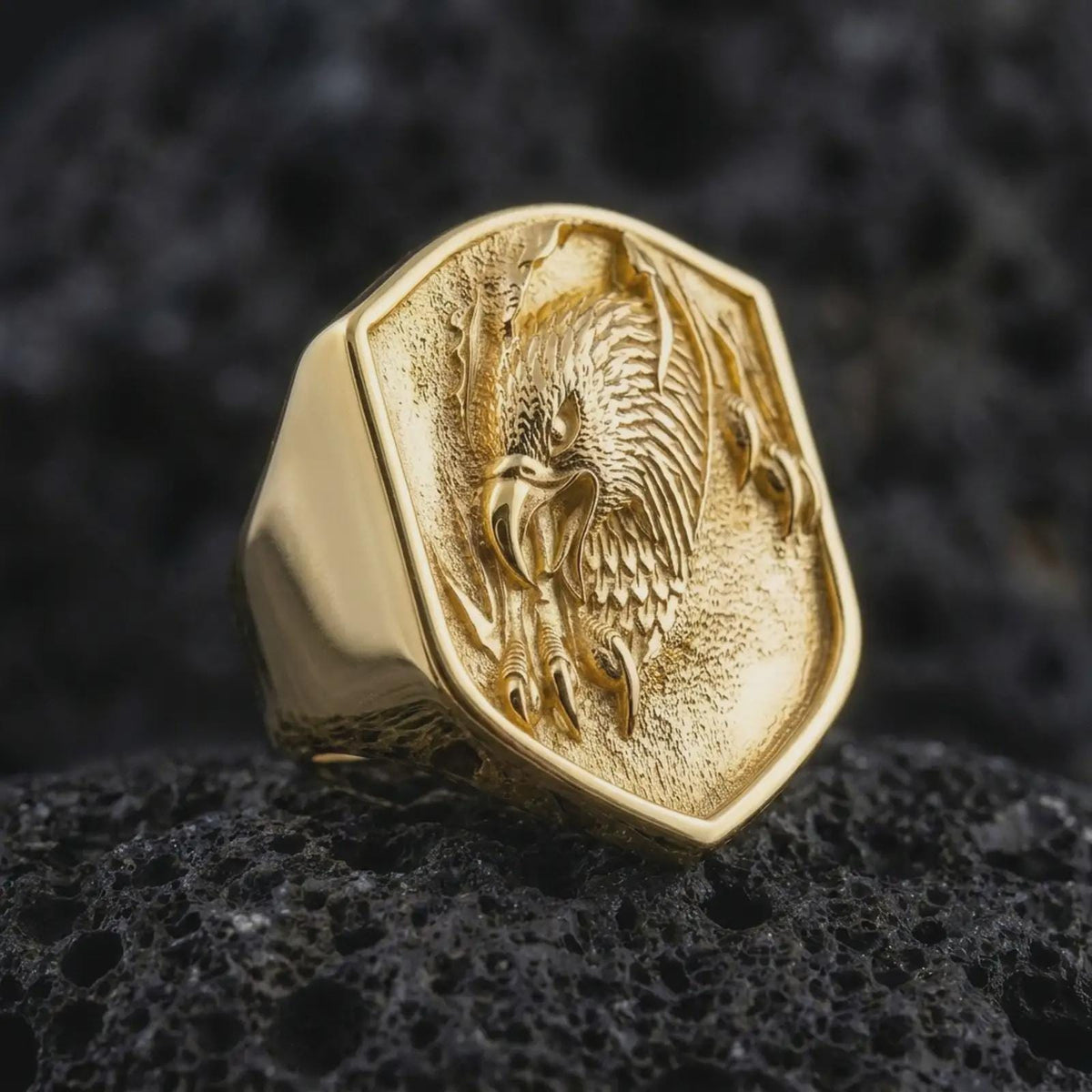 Gold plated 925 sterling silver eagle shield signet ring displayed on textured black volcanic rock, featuring a raised eagle head with an aggressive expression, hooked beak, sharp talons, detailed plumage, and warm gold plated finish.
