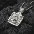 Load image into Gallery viewer, Polished Silver Saint George pendant displayed on black volcanic rock, highlighting the sculpted dragon, Greek inscriptions, and cross-shaped frame.
